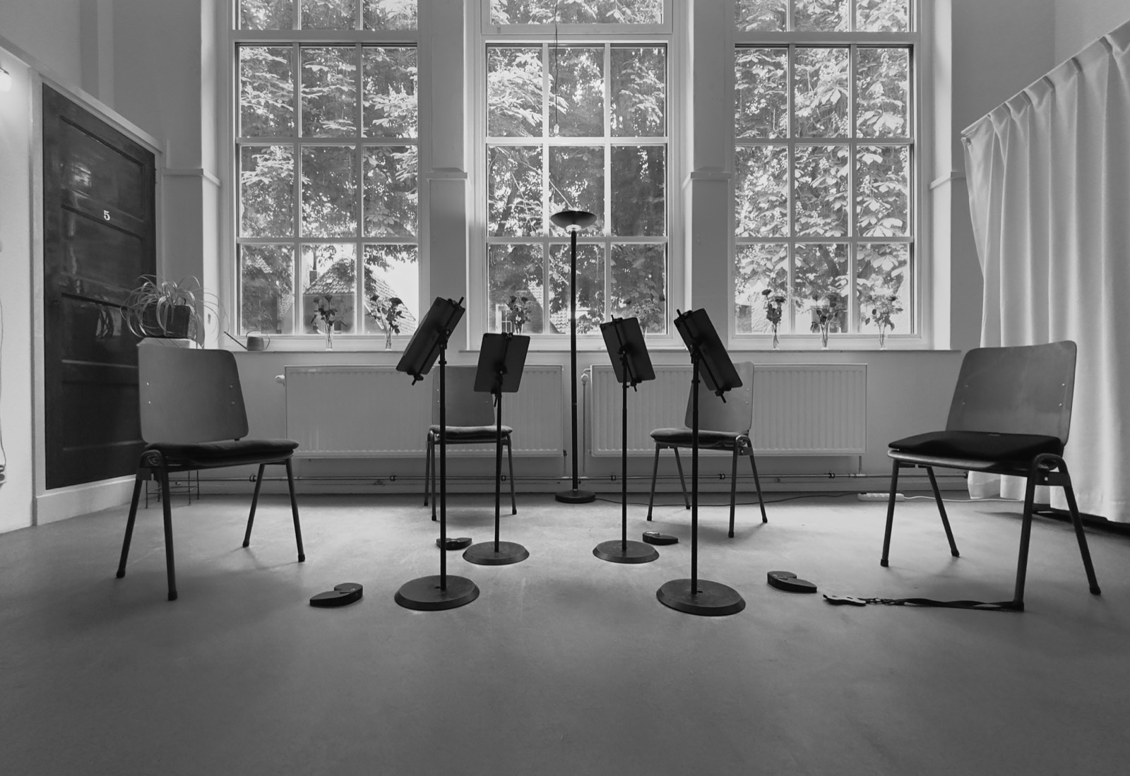 Four empty chairs and music stands arranged in a sunlit room, photographed in black and white.